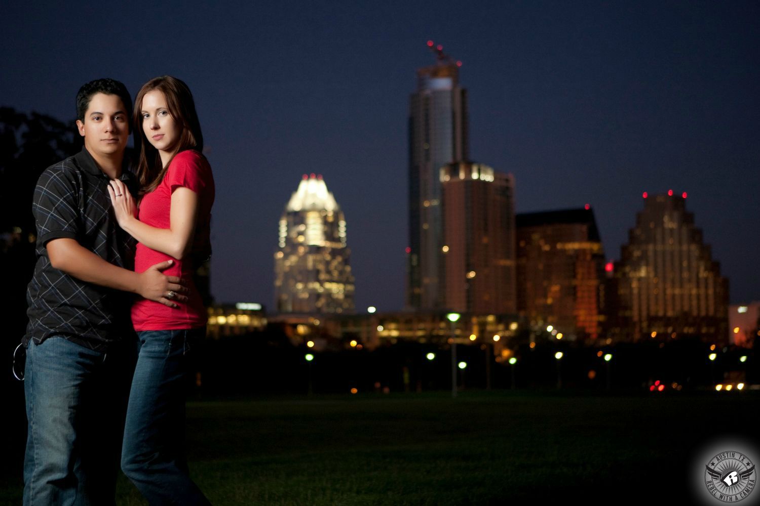 Ginger hair, stoic fair skinned girl in a red low cut blouse and blue jeans stands with her on hand on the chest of a Latino dark haired guy with a navy blue collared crosshatch pattern  shirt and blue jeans at Butler Park in front of the Austin city skyline at night in this vivid engagement image near The Long Center!  